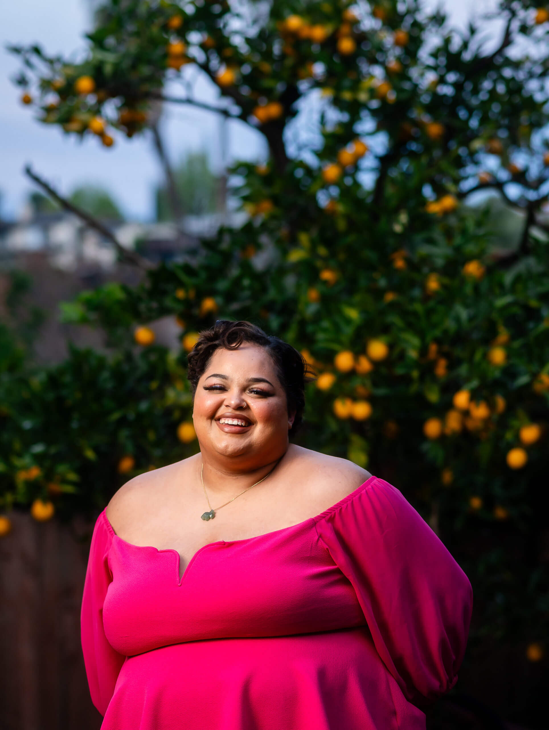 An LGBTQ+ young person in a vivid pink blouse smiles in front of an orange tree.